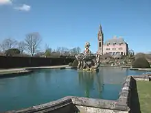 In the foreground is an irregularly-shaped artificial lake containing a statue, behind which are the house and the clock tower