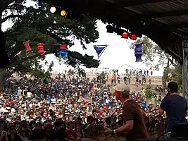 Photo is shot from behind the band looking out to the audience. Two band members are in the foreground at the right. First man (Mikey) is partly turned to his left while playing his guitar. Second man (Huntley) is shown from behind. The other band members are out of view.