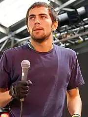 Huntley is shown in an upper body shot. He holds a microphone in his right hand. He wears a purple T-shirt and black gloves. Huntley is looking to his upper right.