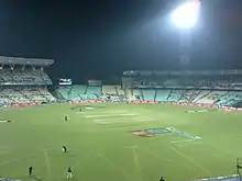 Panoramic view of a cricket ground under floodlights with players occupying the field.