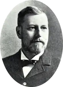 A black and white portrait of a bearded man, wearing a suit with a bowtie