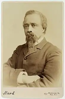 Black-and-white, bust-length portrait photograph of Edward Bannister in a cabinet card mount. He is looking directly at the camera, has his arms crossed, and is wearing a jacket and collared shirt.