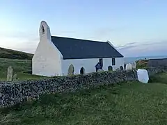 small whitewashed church under slate roof in a walled churchyard with gravestones, and a glimpse of the sea beyond