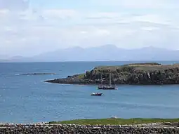 A two-masted yacht and a RIB lie at anchor on a sunny day off  a grassy shore, with low cliffs beyond. A skerry lies further offshore to the left with high hills in the distance.