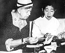 An elderly woman in a hat and a young Asian woman eating lunch using chopsticks