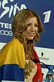 A blond-haired woman sitting at a table wrapped in an oversized Romanian flag.