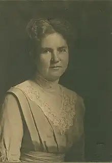 A white woman with greying hair, wearing a dress with a pleated bodice and a lace bib detail