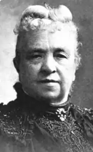 An older white woman with white hair fastened on top of her head and a short curly fringe. She is wearing a dark lace dress with a high collar, and a brooch fastened at her throat.