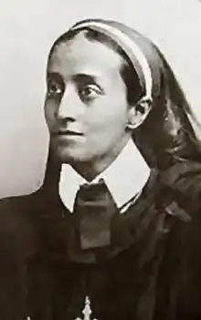 A young South Asian woman, hair dressed back beneath a wide fabric band, wearing a high, stiff, white shirt collar under black robes