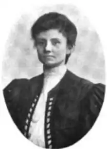 A young white woman with dark hair, wearing a high-collared white blouse under a voluminous dark jacket with a ribbon detail down the front placket