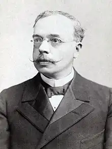 A black and white image of a man in his late middle age. He has formal dress small glasses and a moustache.