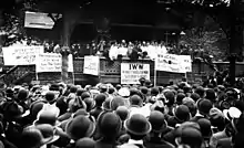 Black and white photograph of a speaker rallying a large crowd. In front of the stage, facing the audience, are several signs, in various languages, displaying demands.