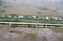 A color photograph taken from the air showing the Everglades bisected by a highway; at the bottom is a sawgrass field flooded with water bordered by a full canal; at the top are some homes and a dry sawgrass field