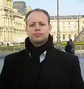 Tenor Ewandro Stenzowski in front of the Louvre museum in Paris, France. He is wearing a black coat and black scarf over a white button down shirt.