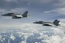 Two aircraft flying high above clouds, transferring fuel through a pipe to which the lower aircraft is connected.