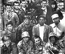 A black and white photo of a group of people, standing around a camera in a wooded area.