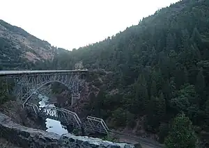 A river flowing between two wooded mountains with a railroad trestle crossing it and a higher road bridge past the trestle