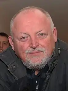 Middle-aged Caucasian male with short hear and facial hair, wearing black jacket and smiling at camera