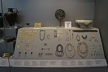 Photograph of a museum cabinet, with various mostly-gold items. A bronze mirror is visible in the top middle.