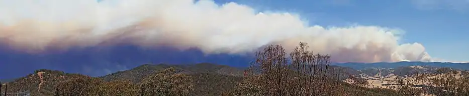 Panorama of a hilly expanse featuring a large smoke trail covering more than half of the visible sky.