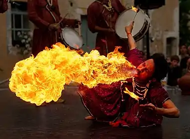 A full-color image shows a high-contrast and quite dramatic scene of a fire breather with a large orange-yellow flame extending from his lips. He wears dark but colorful orange-red clothing.