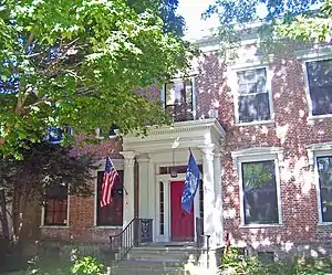 A brick building with a red door behind a small columned porch with the U.S. and New York flags behind a large tree on the left