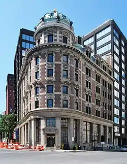 An older, ornate stone and brick building with a small domed green top in a city. There are larger, more modern buildings behind it.