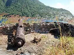 Fort Cachacrou ruins including the remains of a stone wall as well as an old cannon. Below the wall is a body of water, Soufriere Bay.