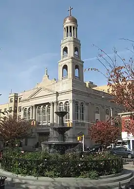 Three-quarter view of a stone church. A three-tiered bell tower topped by a dome top the corner. In the foreground is a black fountain surrounded by shrubbery.