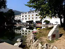 A white building with ornamented windows faces a lake ringed with rock structures. Trees appear around the structure.