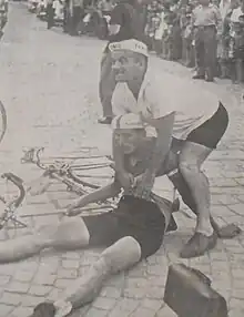 black and white image of Franc Škerlj at approximately 21 years old. He is sitting on the floor next to his bicycle and wearing cycling gear. There is a man standing over him. He looks to have fallen off his bike during a race in front of a crowd