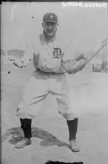 A man in a white baseball uniform with a dark "D" on the front, a dark cap with a white "D", and high socks stands with his arms apart awaiting a throw to his catcher's mitt.