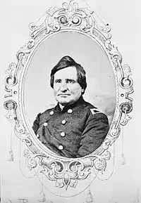 Black and white oval photo shows a clean-shaven man with a mop of hair on top of his head. He wears a dark military uniform with a colonel's shoulder tabs.