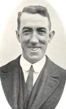 A smiling white man wearing a suit and tie; he is clean-shaven and has short side-parted hair