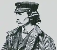 A man in oblique profile with a mustache is wearing a coat and hat looking toward the left.