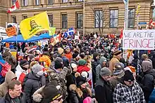 In a dense crowd of people wearing winter clothing in front of a building, several hold flags bearing the National Flag of Canada. Standing shoulder-to-shoulder, the vast majority are not wearing face masks. A large Gadsden flag and a large sign with the words "We are the fringe" are visually prominent.