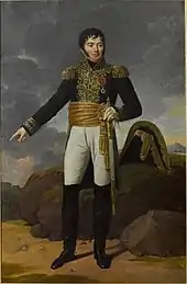 Formal full-length portrait of a Tharreau in uniform, in a rocky landscape. He is a short, solidly built man with a round face and alert dark eyes. His pale skin contrasts with his black curly hair and heavy cheek whiskers. He looks out of the picture to the left while gesturing backward with his right hand. He holds a sword in his left hand.