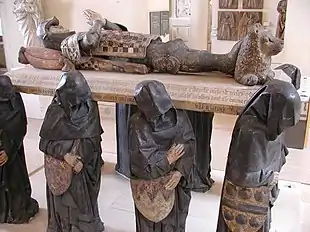 A large sculpture of six life-sized black-cloaked men, their faces obscured by their hoods, carrying a slab upon which lies the supine effigy of a knight, with hands folded together in prayer. His head rests on a pillow, and his feet on a small reclining lion.