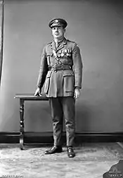 A full-length portrait of a man in military uniform wearing medals. His hand is resting atop a small table.