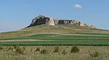 Steep-sided rocky bluff surrounded by grassland; green fields in foreground