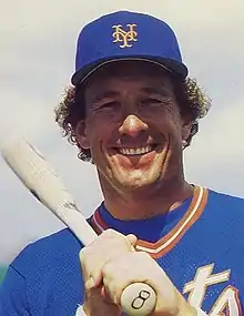 A man in a New York Mets  blue baseball jersey and cap with a bat resting on his shoulder