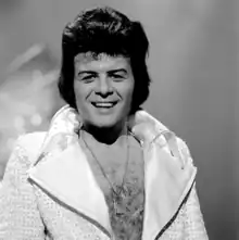 A black-haired man with a hairy chest, wearing a shiny jacket open to the waist, with large lapels, smiles towards the camera