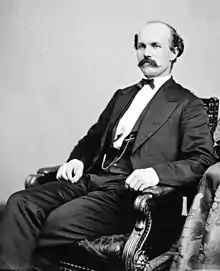 Black and white photo of a balding man with a moustache sitting in a chair. He wears a dark suit and pants with a white vest.