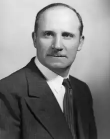 A middle-aged man in a suit, with receding hair and a neat moustache