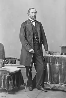 Fair-skinned man with dark hair and beard, standing in a formal portrait pose, wearing mid-Victorian clothes and holding a cane