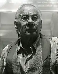 Shoulder-high portrait of a man in his seventies, with gray hair, wearing glasses a striped shirt and a vest made of patchwork suitings