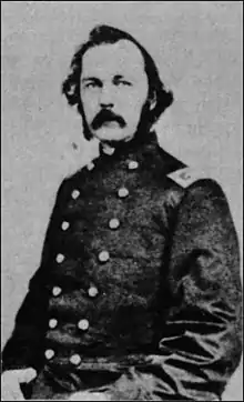 Black and gray photo of a man with a moustache and long sideburns. He wears a dark military uniform with two rows of buttons.