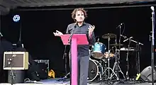 Sam stands behind a pink lecturn framed by an open shipping container with a drum kit and microphones in it. He is talking and has a grey shirt, short curly hair and pale skin.