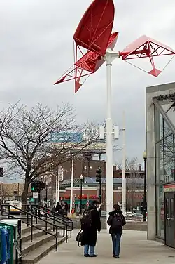 A slender white pole supporting three red, fan-like metal structures all on the same plane but twisted at different angles.