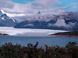 A glacier flows down to a bright blue lake surrounded by snow-capped mountains